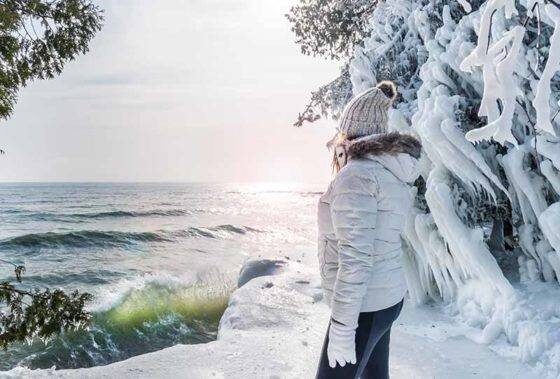 Photo of Woman in white winter coat looking out at ocean from a snowy winter landscape at Cave Point Park, Door County, WI